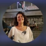 Ανθρώπινη γυναίκα χαμογελά σε εκδήλωση Cosmosyndesis, με φόντο την πόλη και φωτισμό.